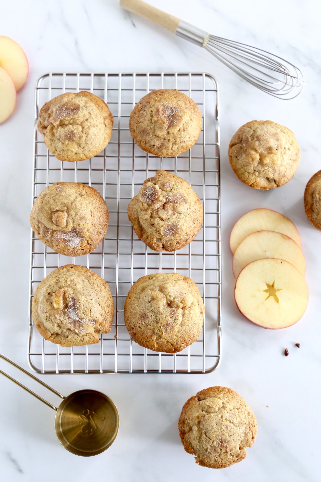 A cooling rack with muffins stacked on top next to a whisk and sliced apples. 