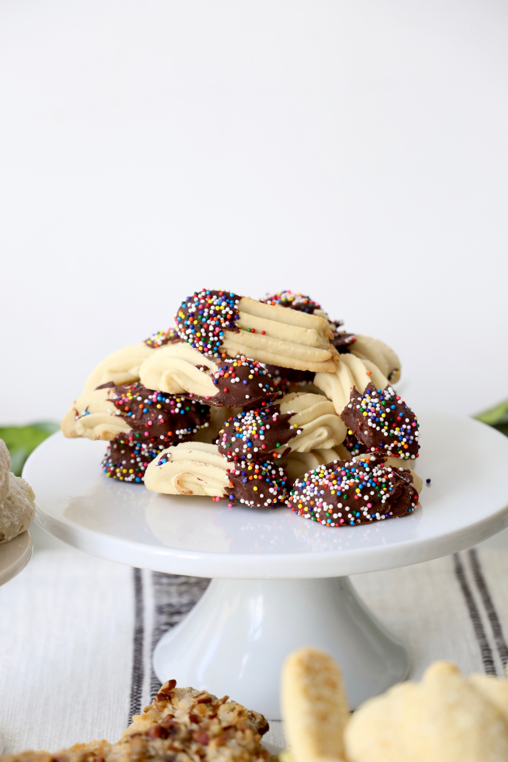 A white cake stand filled with cookies dipped in chocolate and decorated with sprinkles.
