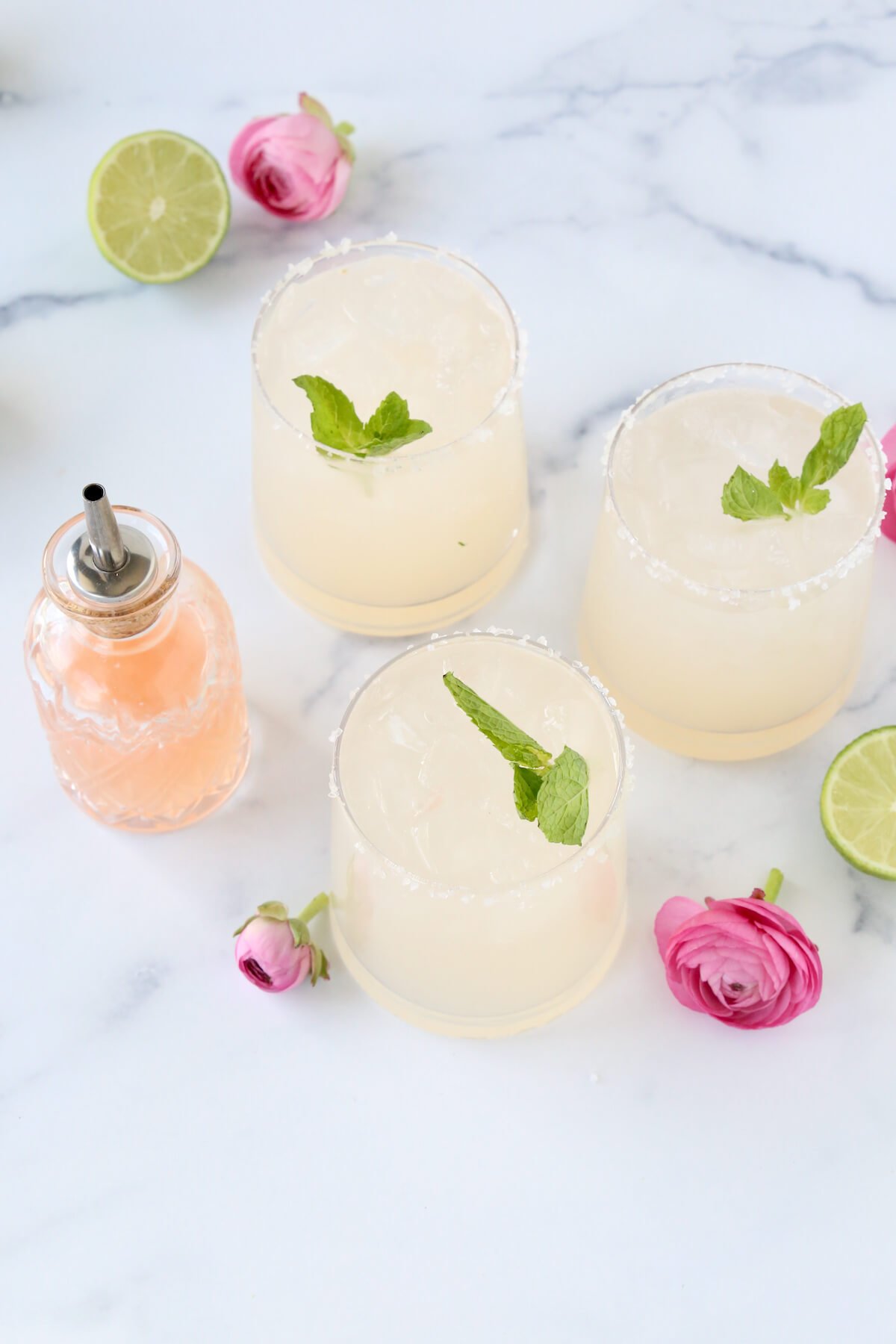 Three glasses with grapefruit, lime juice and tequila mixed together with a fresh mint leave on top next to pink flowers.