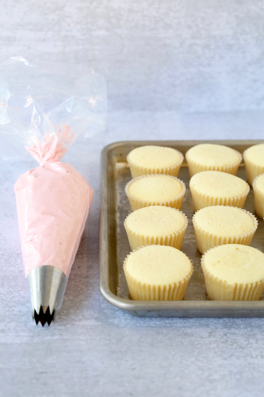 A baking sheet lined with cupcakes and a piping bag filled with pink frosting sitting next to it.  