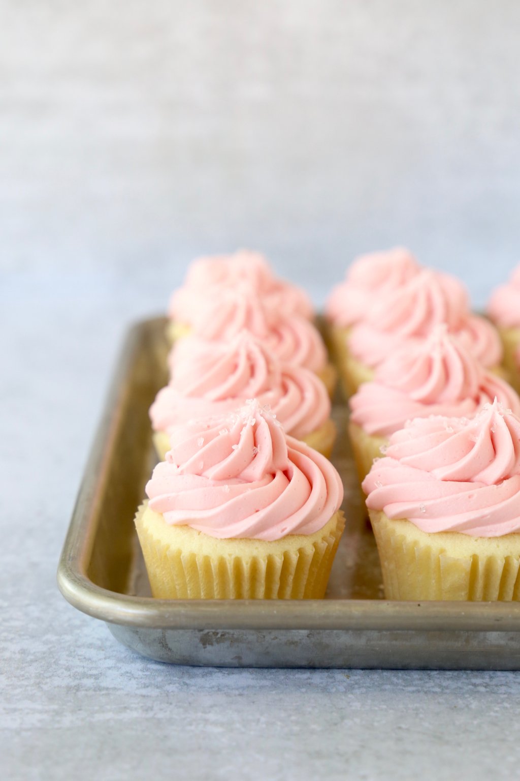 A baking sheet filled with vanilla cupcakes with pink frosting.