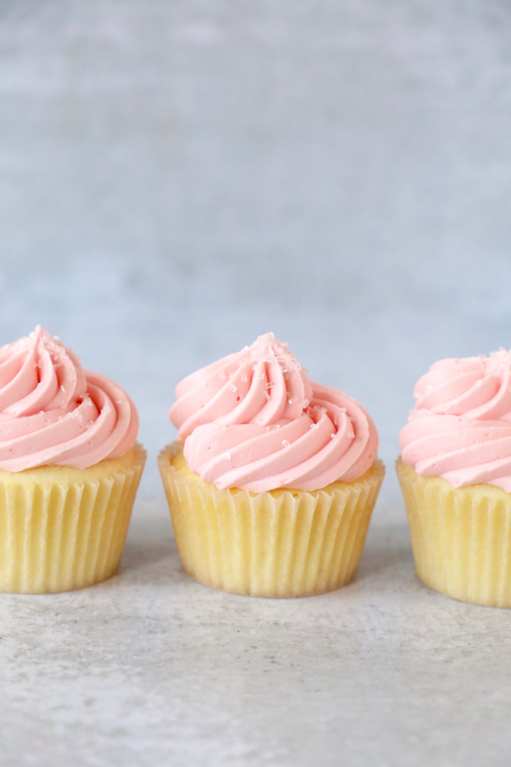 Three vanilla cupcakes with pink frosting and sugar crystals.  