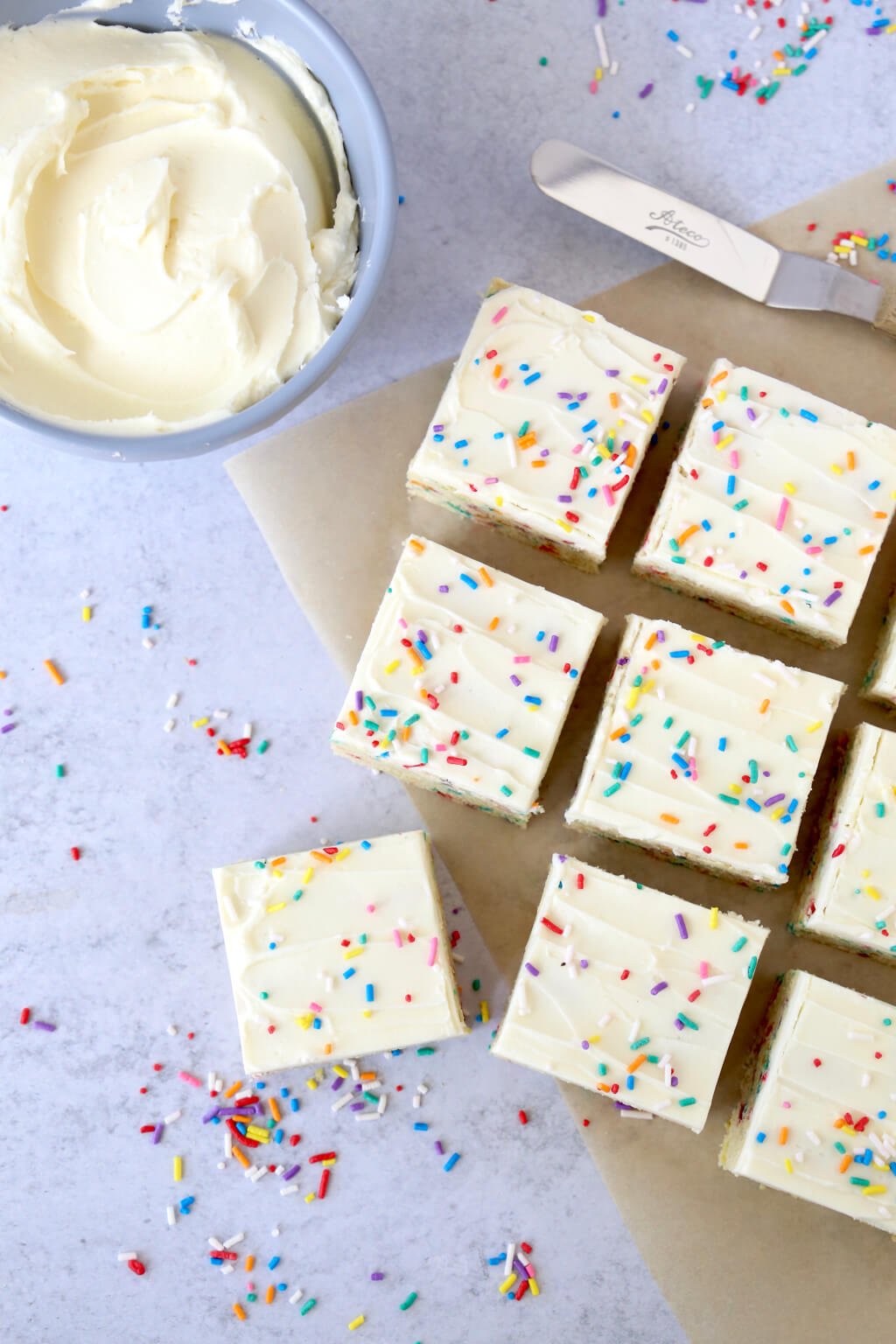 Sugar cookie bars covered in rainbow sprinkles next to a bowl of buttercream frosting