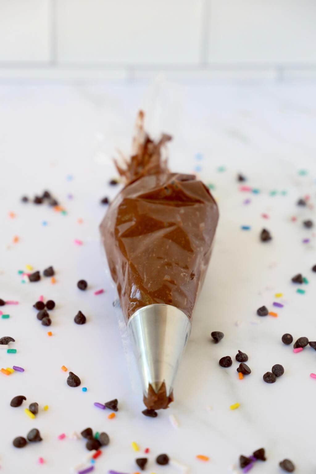 A piping bag filled with chocolate buttercream frosting, surrounded by chocolate chips and rainbow sprinkles
