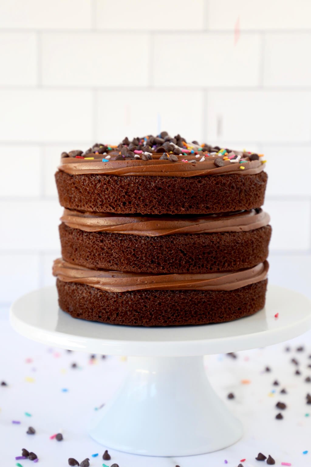 Classic chocolate layer cake topped with sprinkles and chocolate chips on a white cake stand