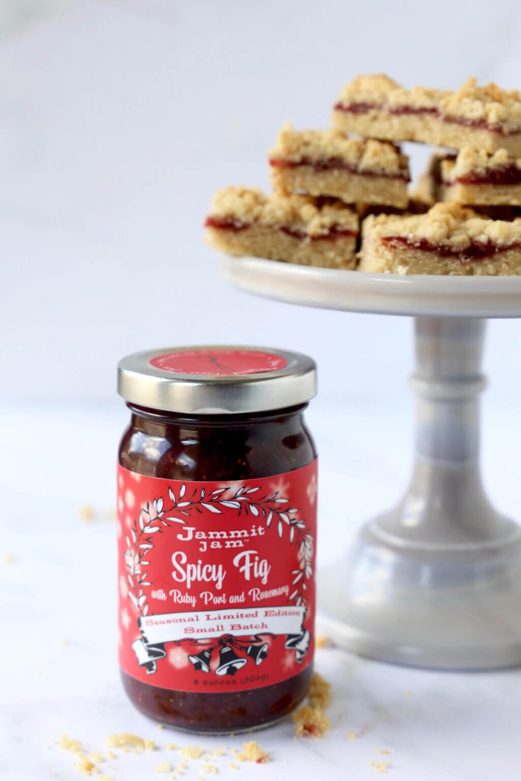 a jar of jammit jam next to a cake stand with a stack of jam shortbread bars 