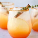 a close up of three glasses of a red orange colored cocktail with a cinnamon stick and thyme leave in each glass