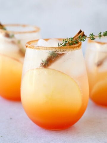 a close up of three glasses of a red orange colored cocktail with a cinnamon stick and thyme leave in each glass