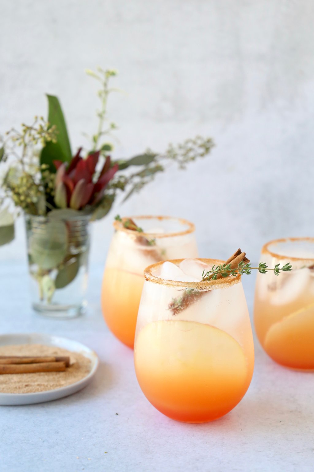 three glasses filled with an orange/red cocktail with a cinnamon stick and thyme leaf sitting next to a small floral arrangement and a dish with cinnamon sugar