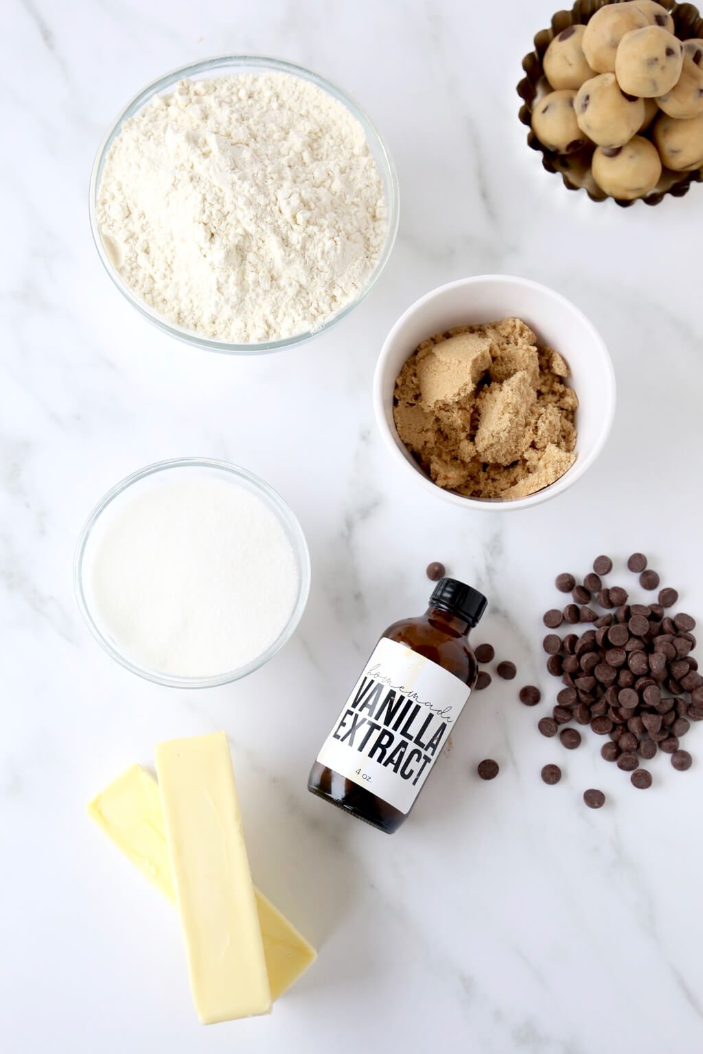 All the ingredients for cookie dough balls. A bowl of flour, a bowl of brown sugar, 2 sticks of butter, vanilla extract, a bowl of heavy cream, and pile of chocolate chips.