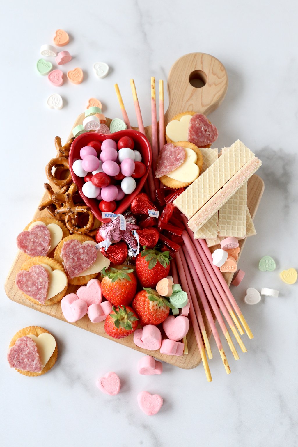 a wood cutting board filling with chocolate, marshmallows, strawberries, wafer cookies and salami and cheese crackers 