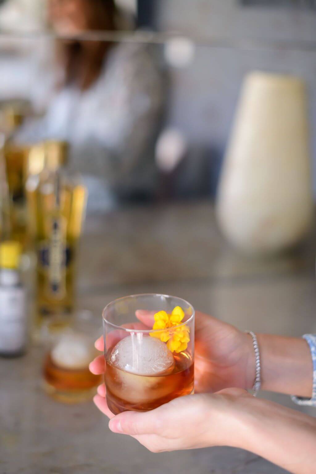 a women holding a clear glass with a big ice ball filled with elderflower, bourbon, bitters and an edible flower