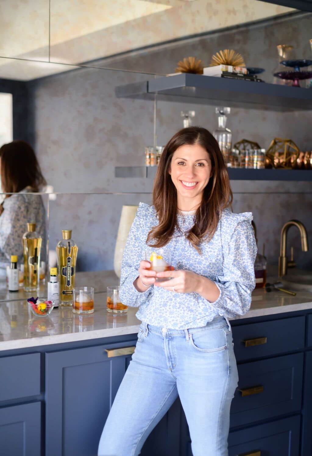 a women holding a clear glass with a big ice ball filled with elderflower, bourbon, bitters and an edible flower