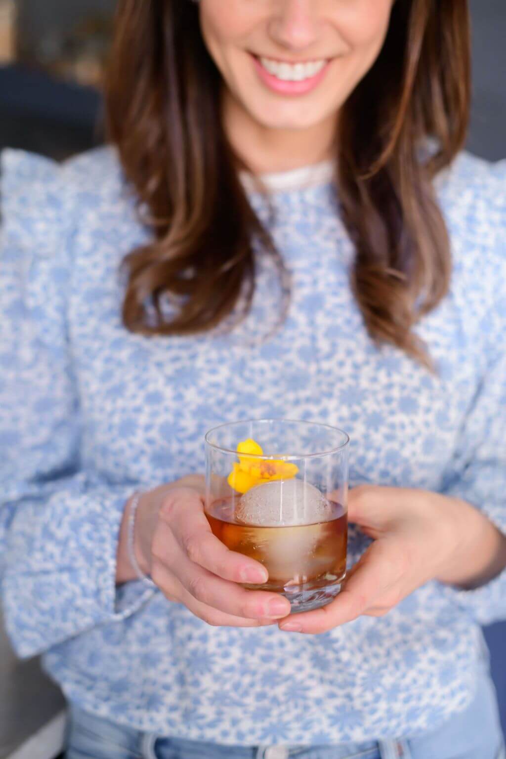 a women holding a clear glass with a big ice ball filled with elderflower, bourbon, bitters and an edible flower