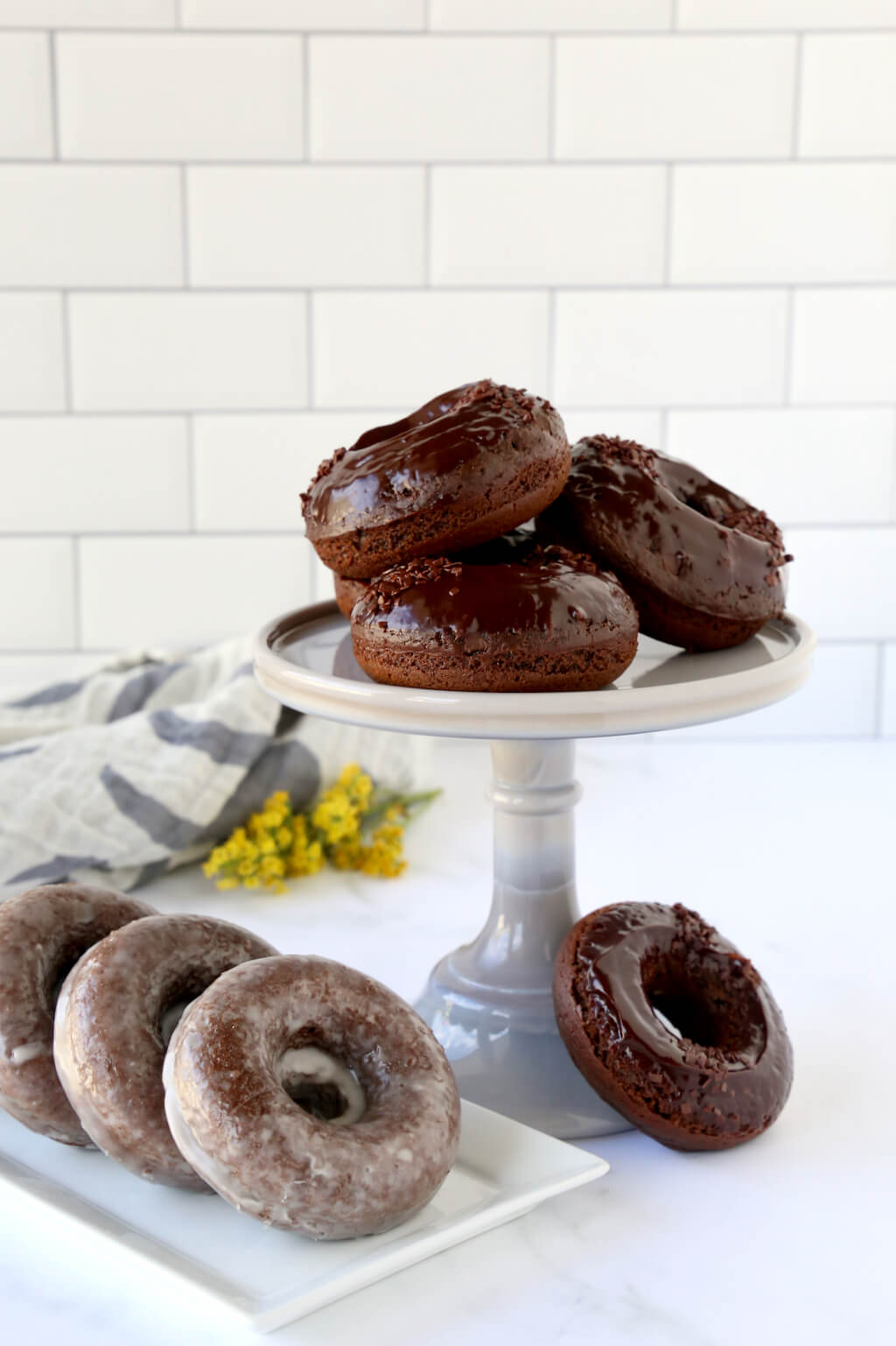 a cake stand stacked with chocolate donuts and a platter of glazed donuts next to it