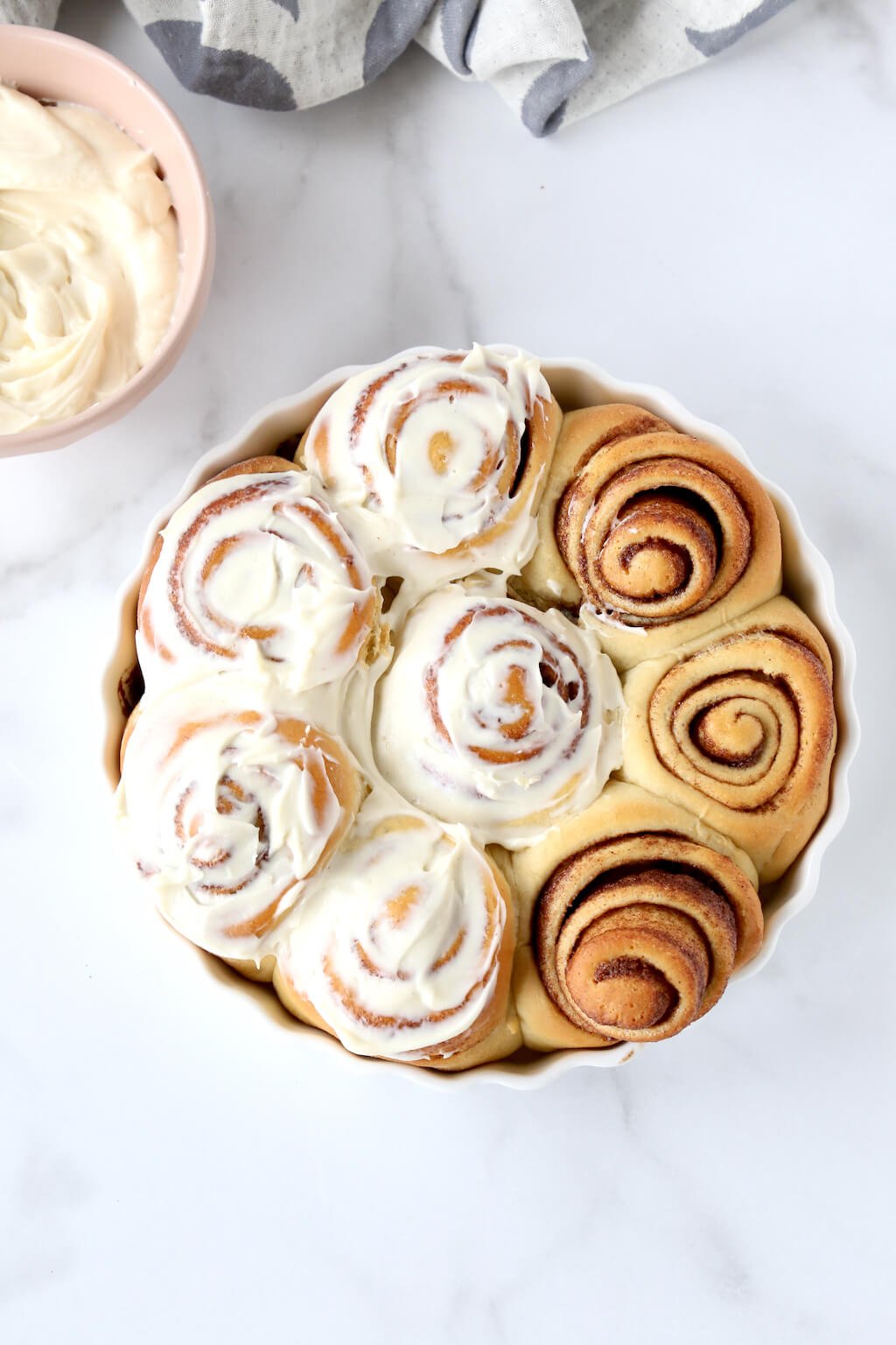a round dish filled with baked cinnamon rolls half glazed with cream cheese frosting 