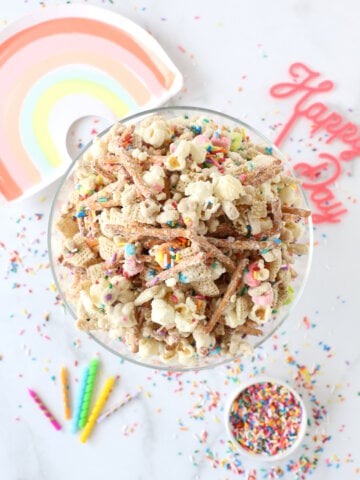 a bowl of snack mix surrounded by sprinkles, a rainbow plate, candles and sprinkles.