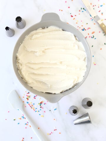 a bowl of white frosting surrounded by sprinkles, piping tips and spatulas.