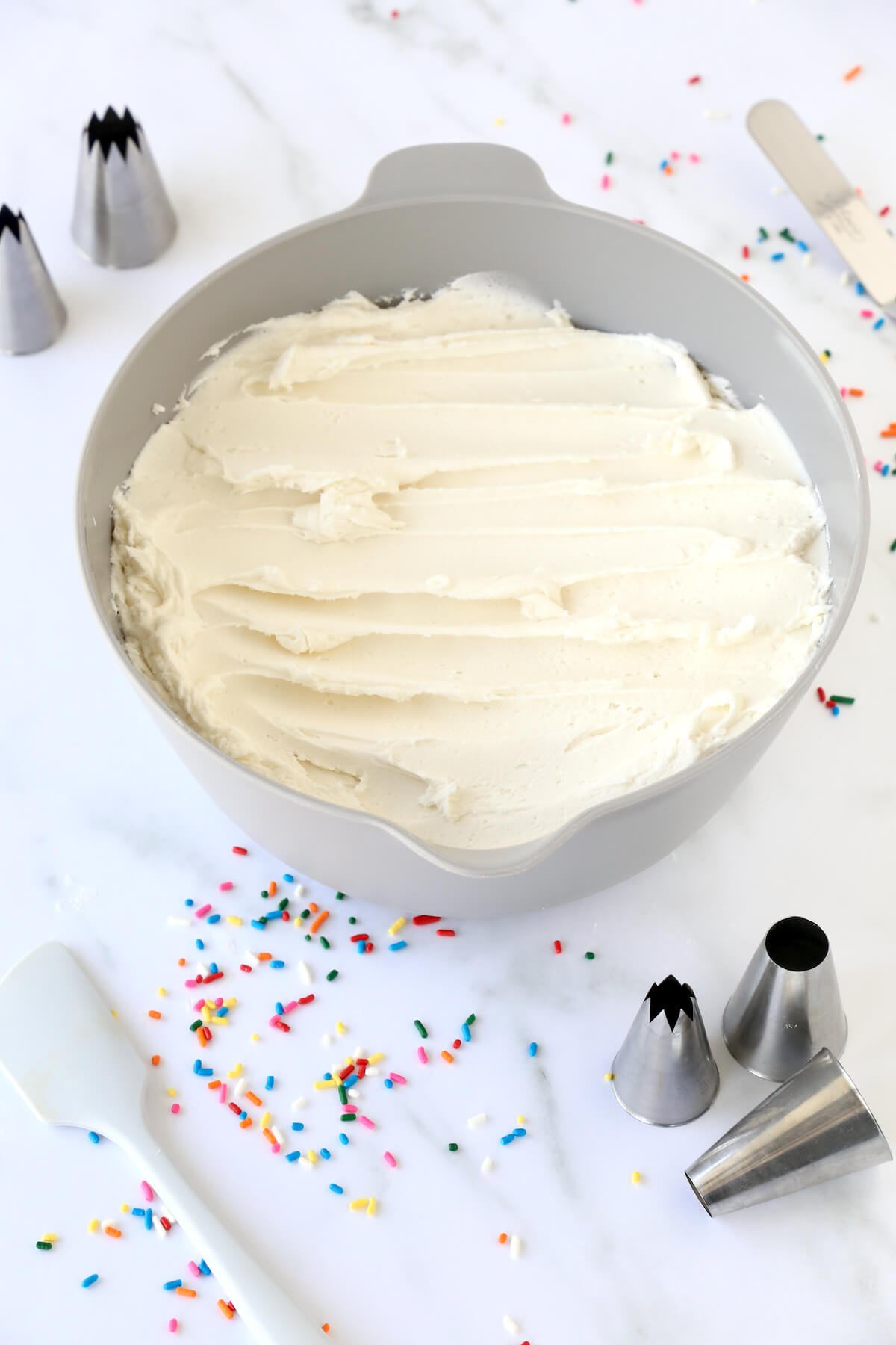 a bowl of white frosting surrounded by sprinkles, piping tips and spatulas.  