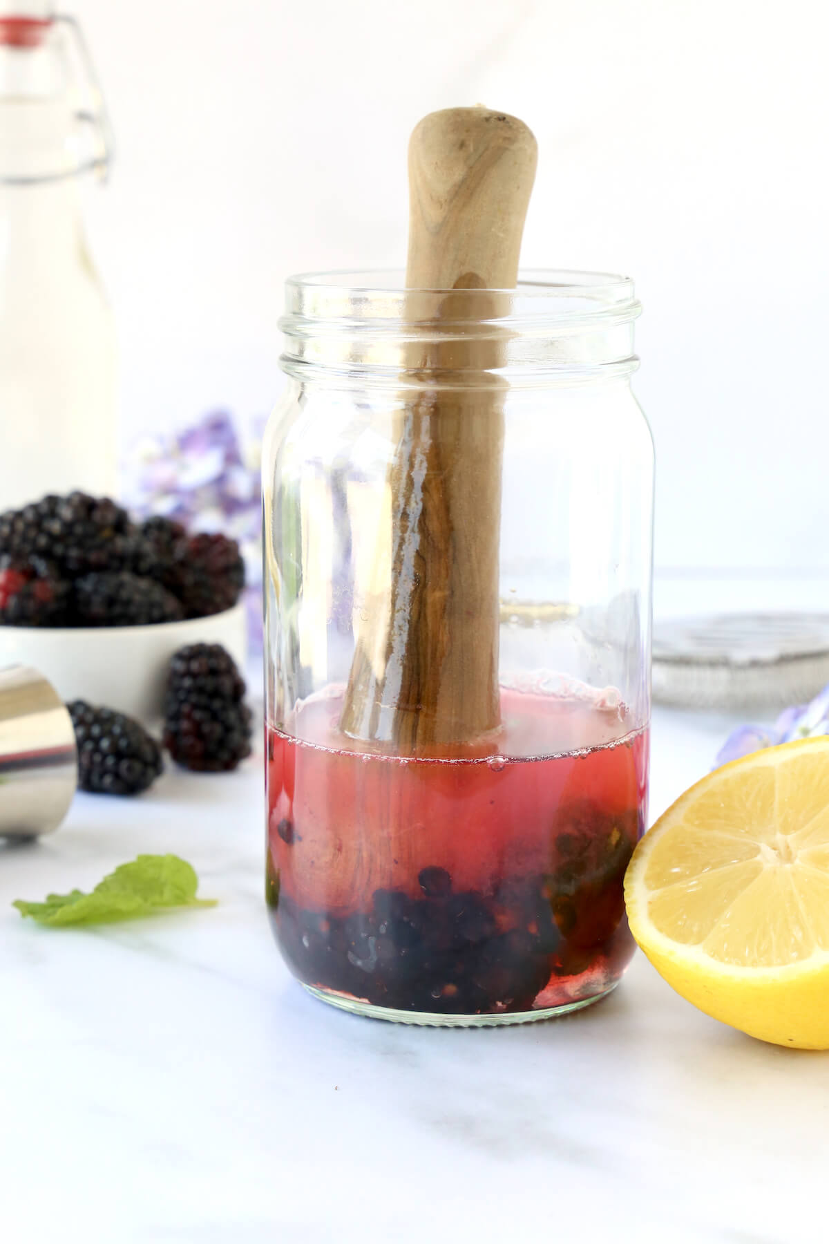 A glass jar filled with blackberries, mint leaves, tequila, lemon juice and simple syrup getting mashed.  