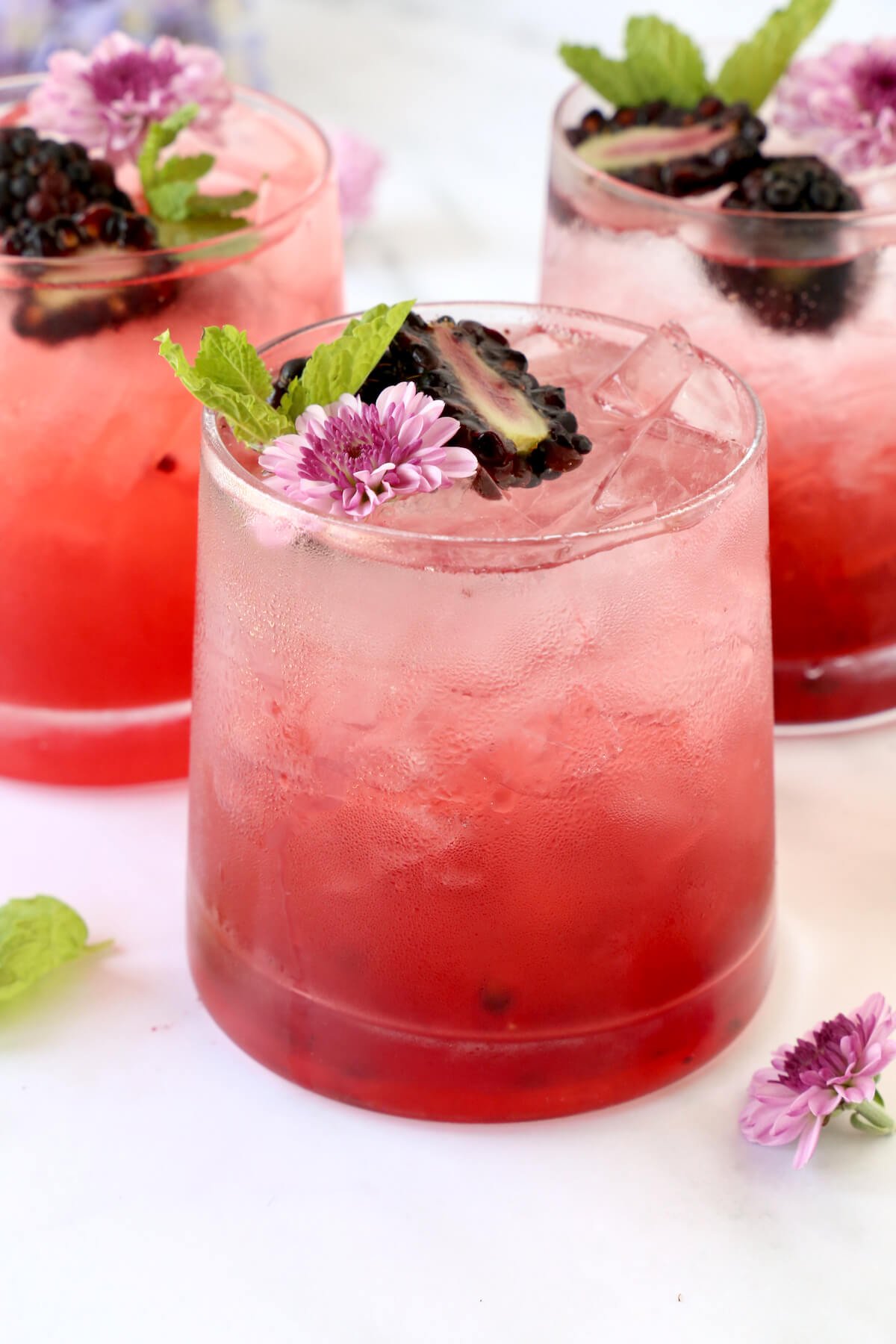 Three clear glasses filled with a red drink, blackberries, mint and a flower.