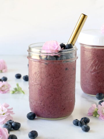 A clear glass jar filled with blueberry smoothie, fresh blueberries and a fresh flower and a gold straw.