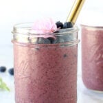 A clear glass jar filled with blueberry smoothie, fresh blueberries and a fresh flower and a gold straw.