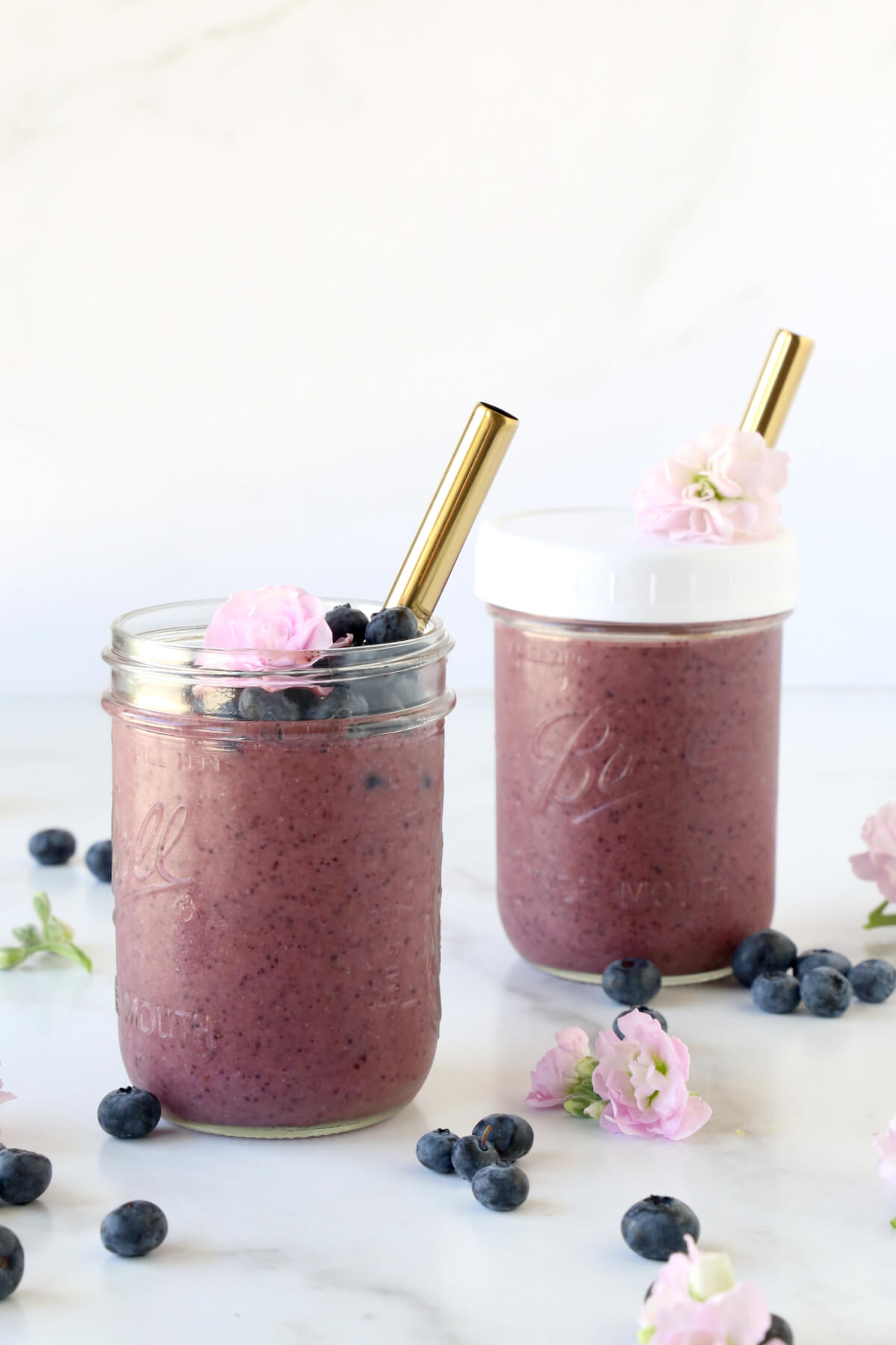 Two clear glass jars filled with blueberry smoothie, fresh blueberries and a fresh flower and a gold straw.