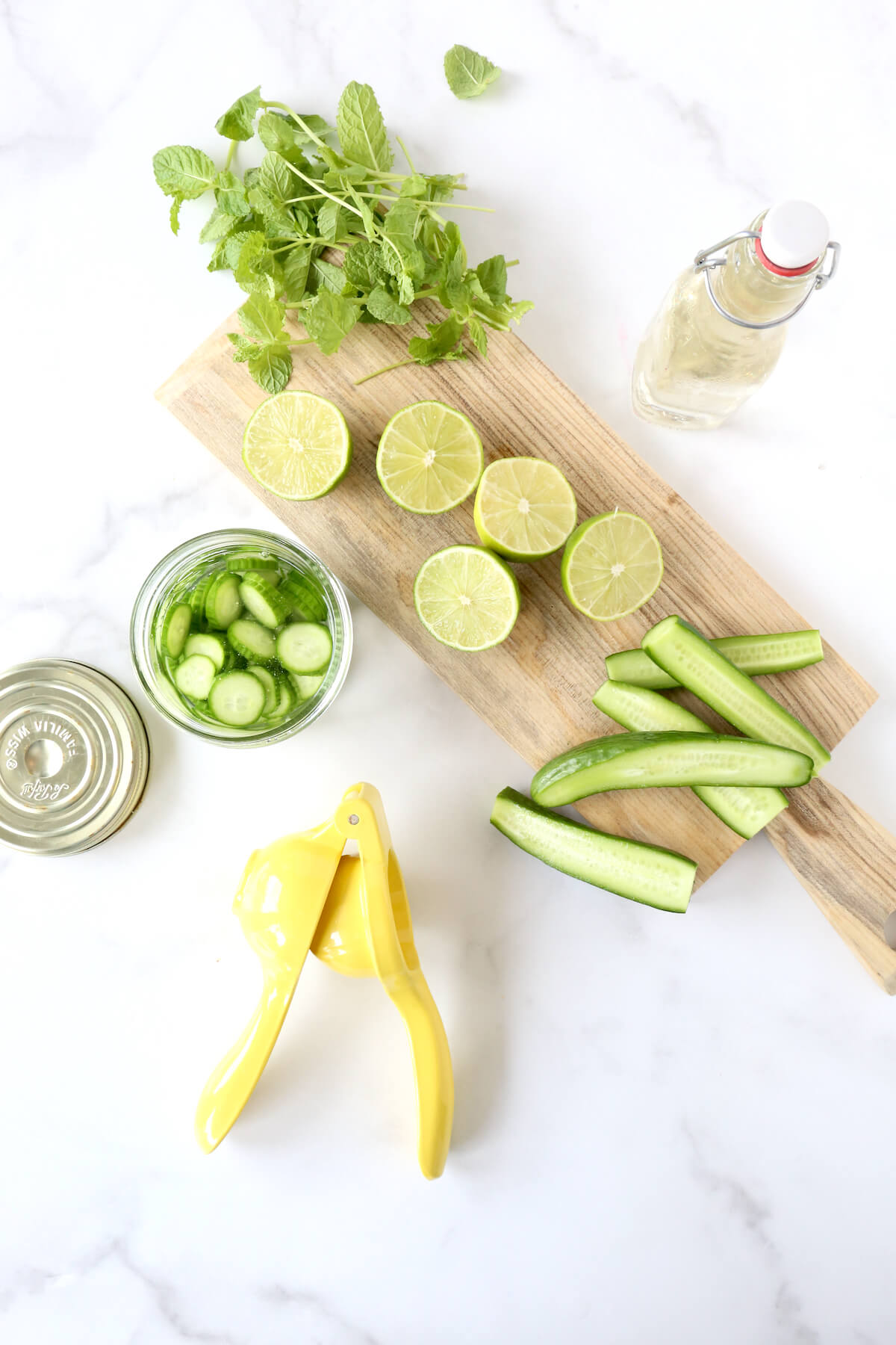 A wood board with sliced limes, cucumbers, mint a juicer and a jar of simple syrup.  