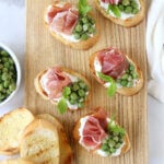 A wood board topped with toasted bread, peas and prosciutto slices.