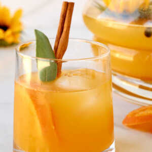 A clear glass filled with an orange liquid, orange slices, cinnamon stick and sage.
