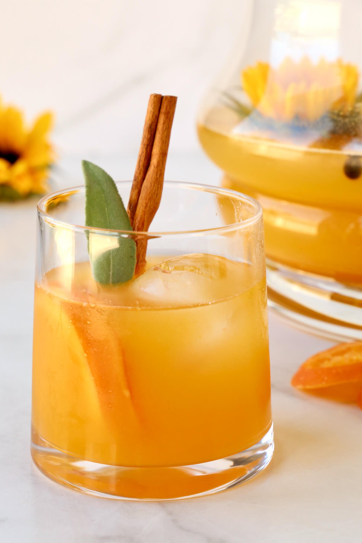 A clear glass filled with an orange liquid, orange slices, cinnamon stick and sage. 