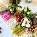 A large platter filled with grape leaves, pickles, hummus, penne, bread, cheese and herbs.