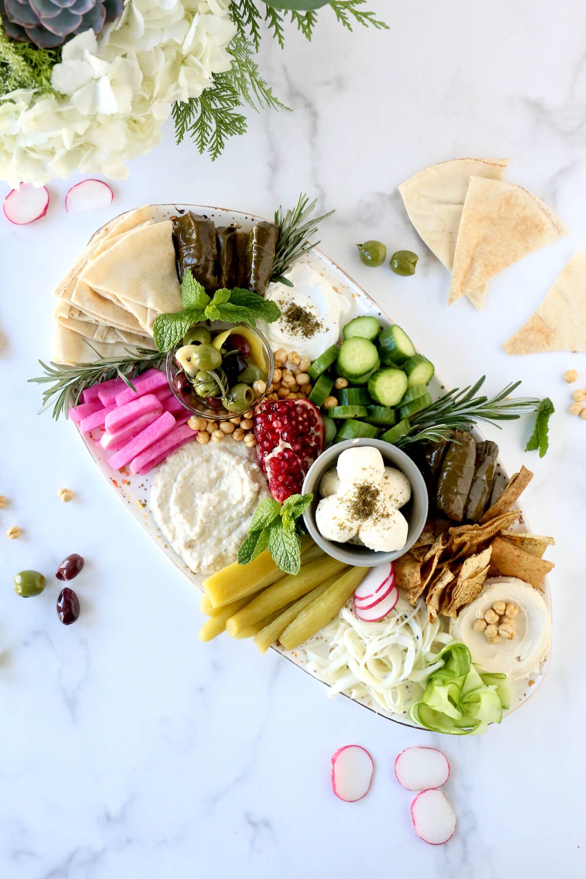 A large platter filled with grape leaves, pickles, hummus, penne, bread, cheese and herbs.