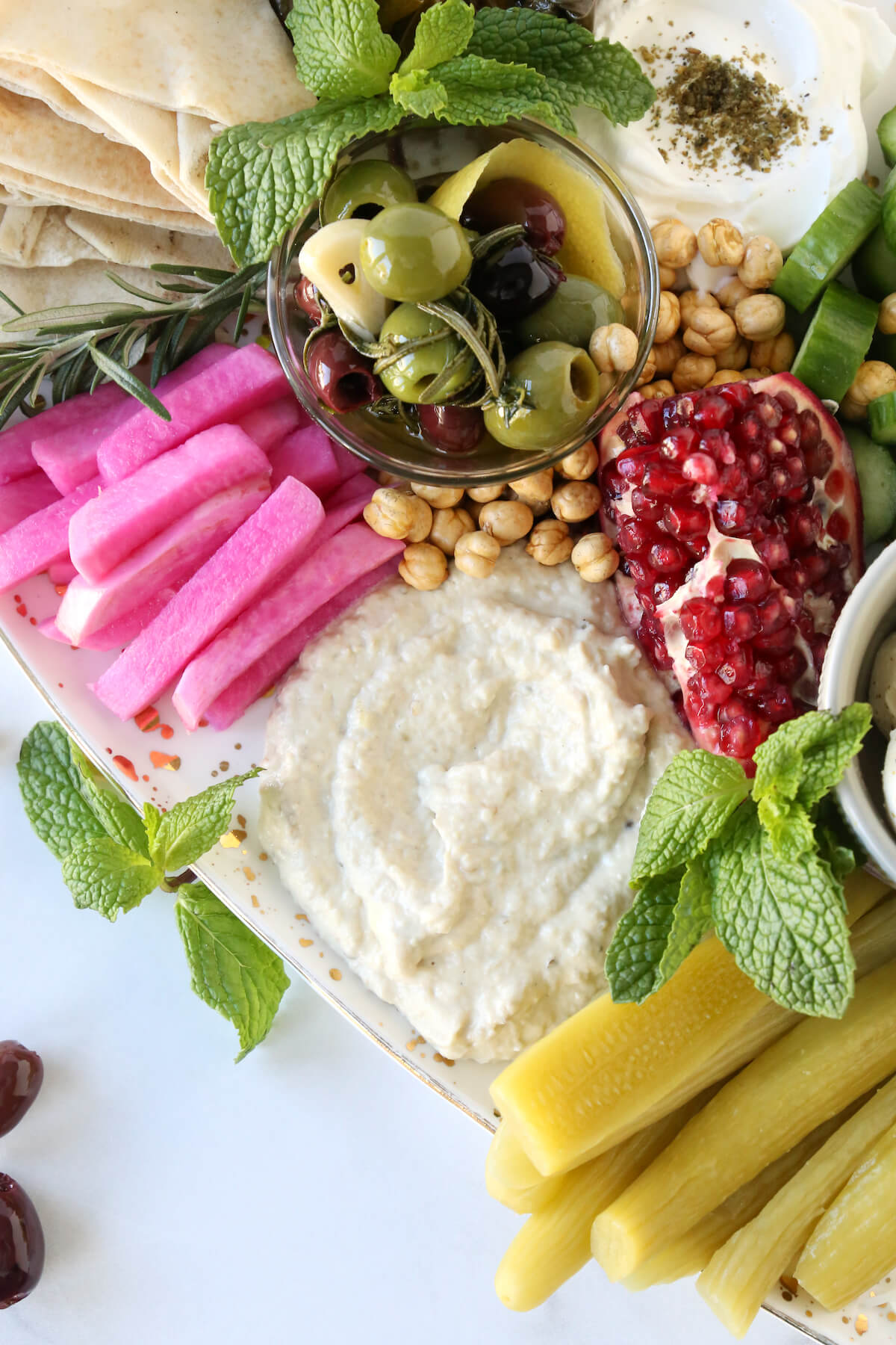 A large platter filled with grape leaves, pickles, hummus, penne, bread, cheese and herbs. 
