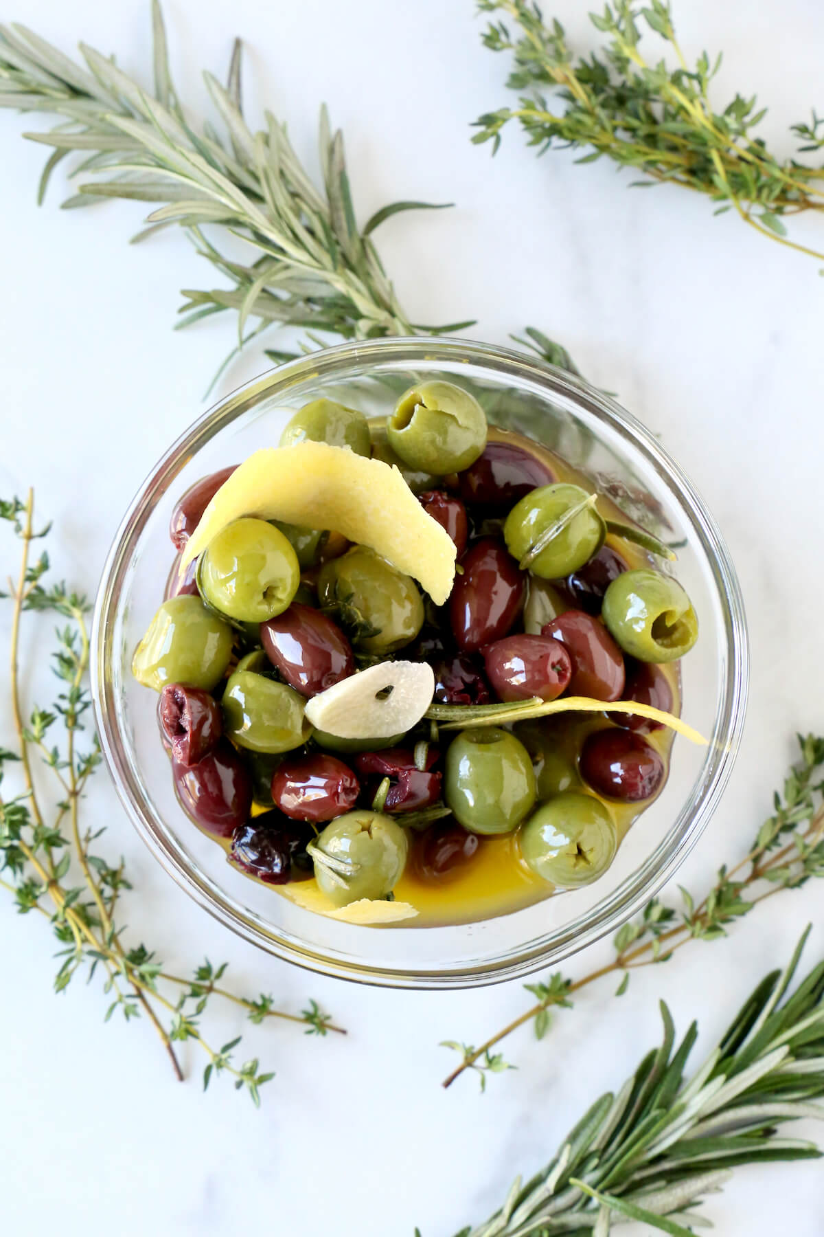 A glass bowl filled with olives, lemon peel and garlic surrounded by fresh rosemary sprigs and thyme.