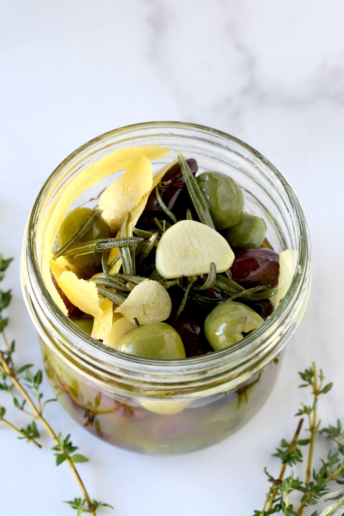 A glass jar filled with olives, lemon peel, garlic and herbs. 