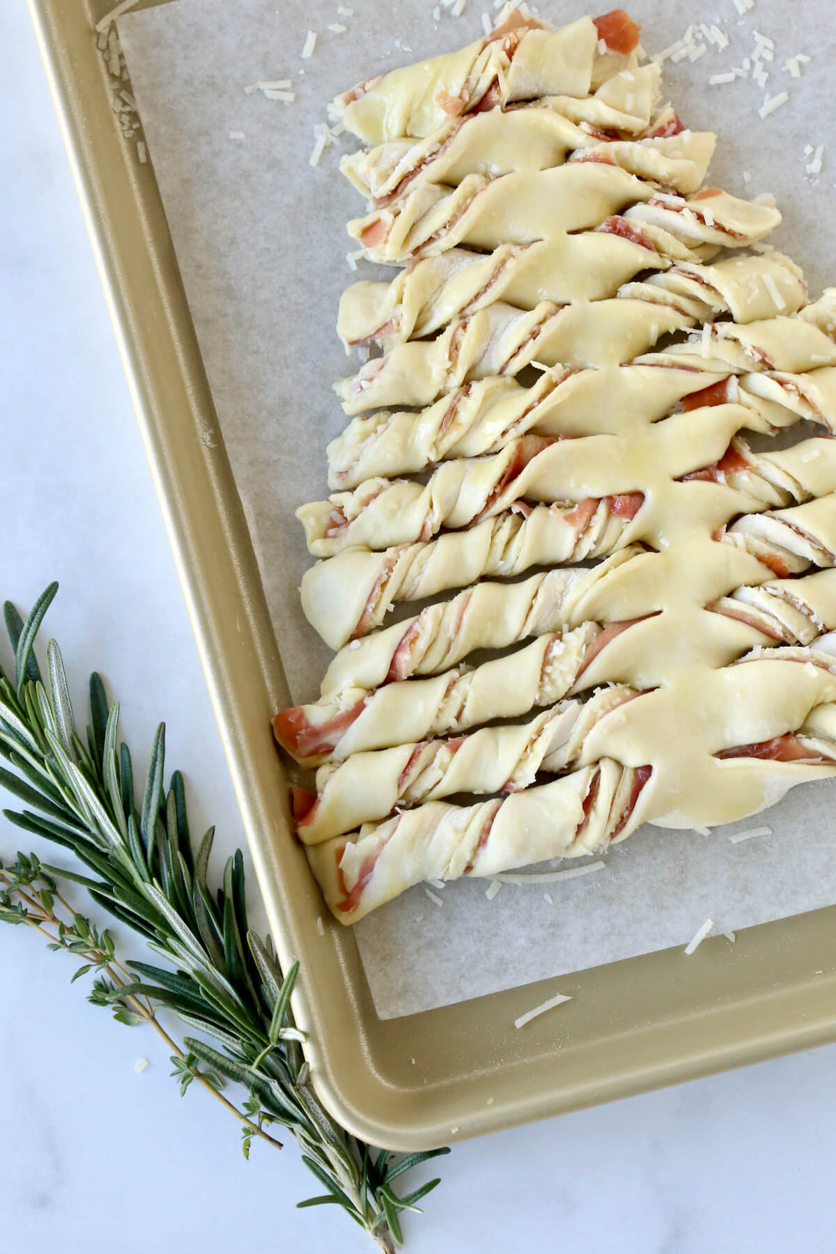 A gold baking sheet with an unbaked tree shaped puff pastry with fresh herbs next to it.