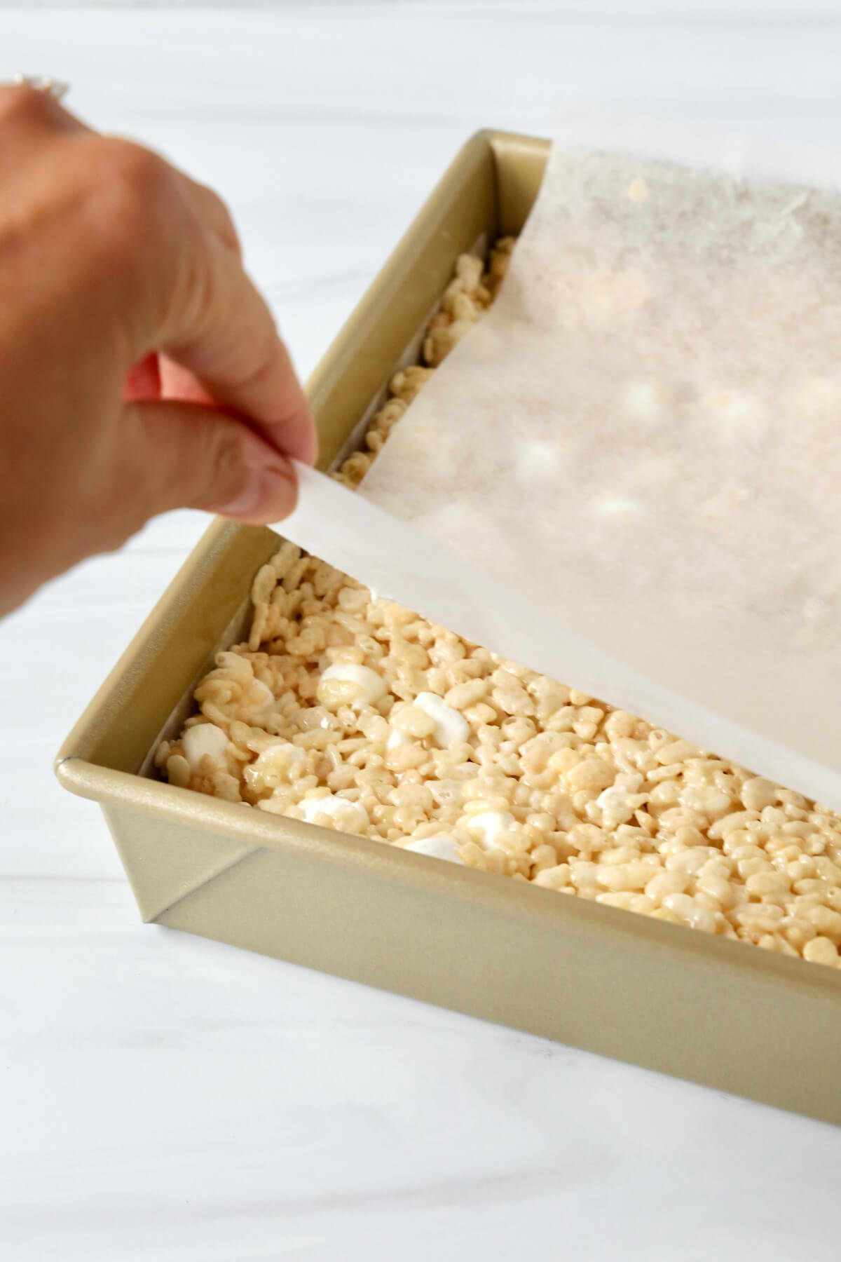 A square gold cake pan with rice krispie cereal pressed down and a hand holding parchment paper on top. 