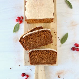 A sliced bread loaf on a wood board with white glaze on top and fresh cranberries and sage leaf.