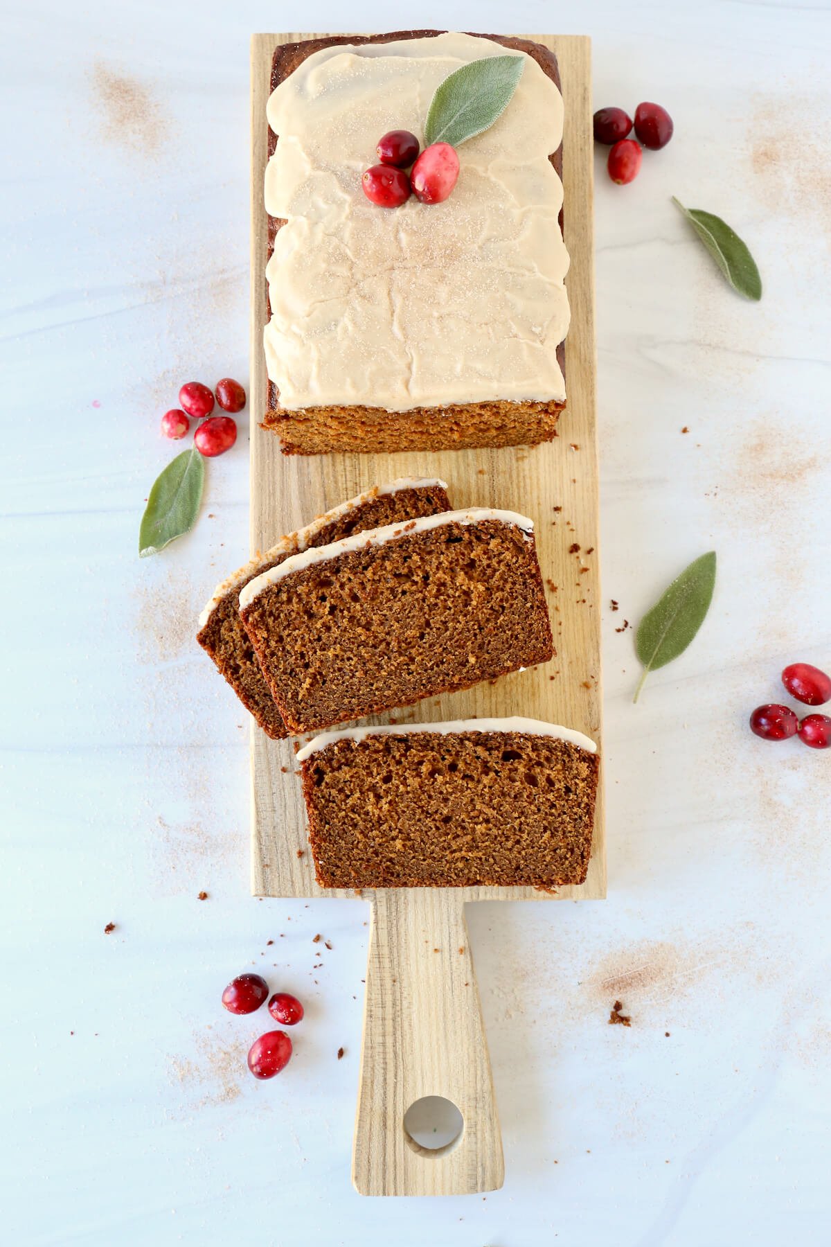 A sliced bread loaf on a wood board with white glaze on top and fresh cranberries and sage leaf. 