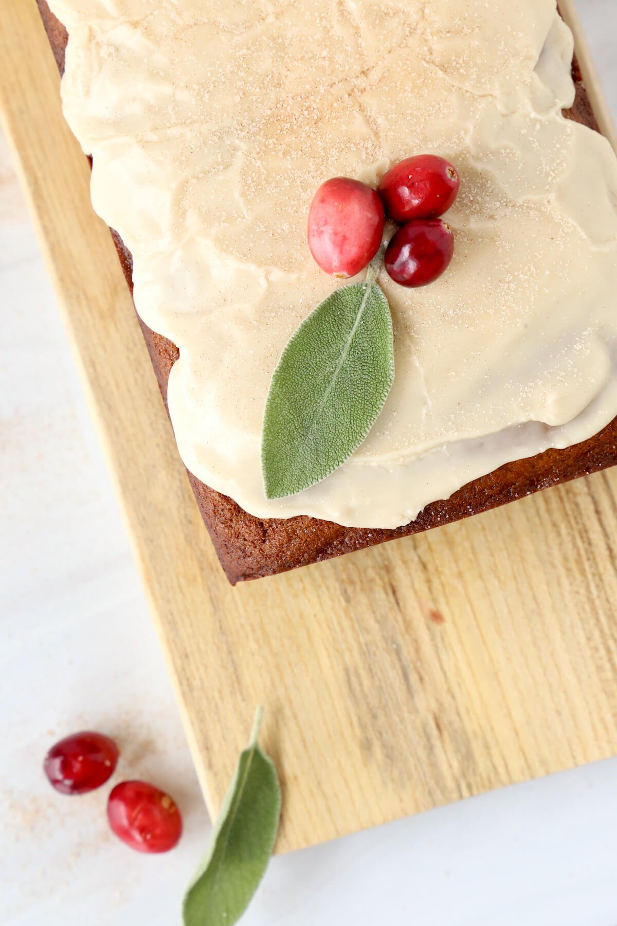 A bread loaf on a wood board with white glaze on top and fresh cranberries and sage leaf.