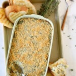 A green baking dish with crushed ritz crackers on the top, next to grilled bread, two spoons and fresh herbs.