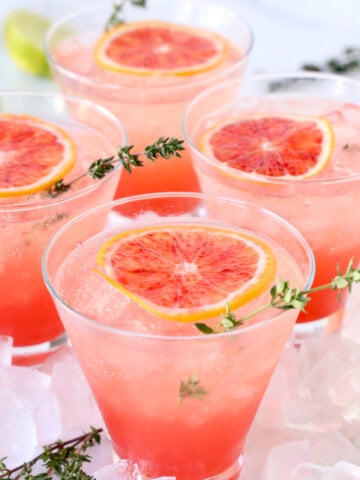 Four glasses or orange liquid with a slice of blood orange and a thyme sprig.