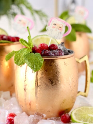 A copper mug filled with ice, cranberries, mint leaves, santa stick and lime.