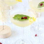 Three coupe glasses filled with a clear liquid, basil leaf, pomegranate seeds and a lemon rind curl.