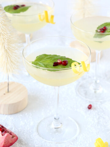 Three coupe glasses filled with a clear liquid, basil leaf, pomegranate seeds and a lemon rind curl.