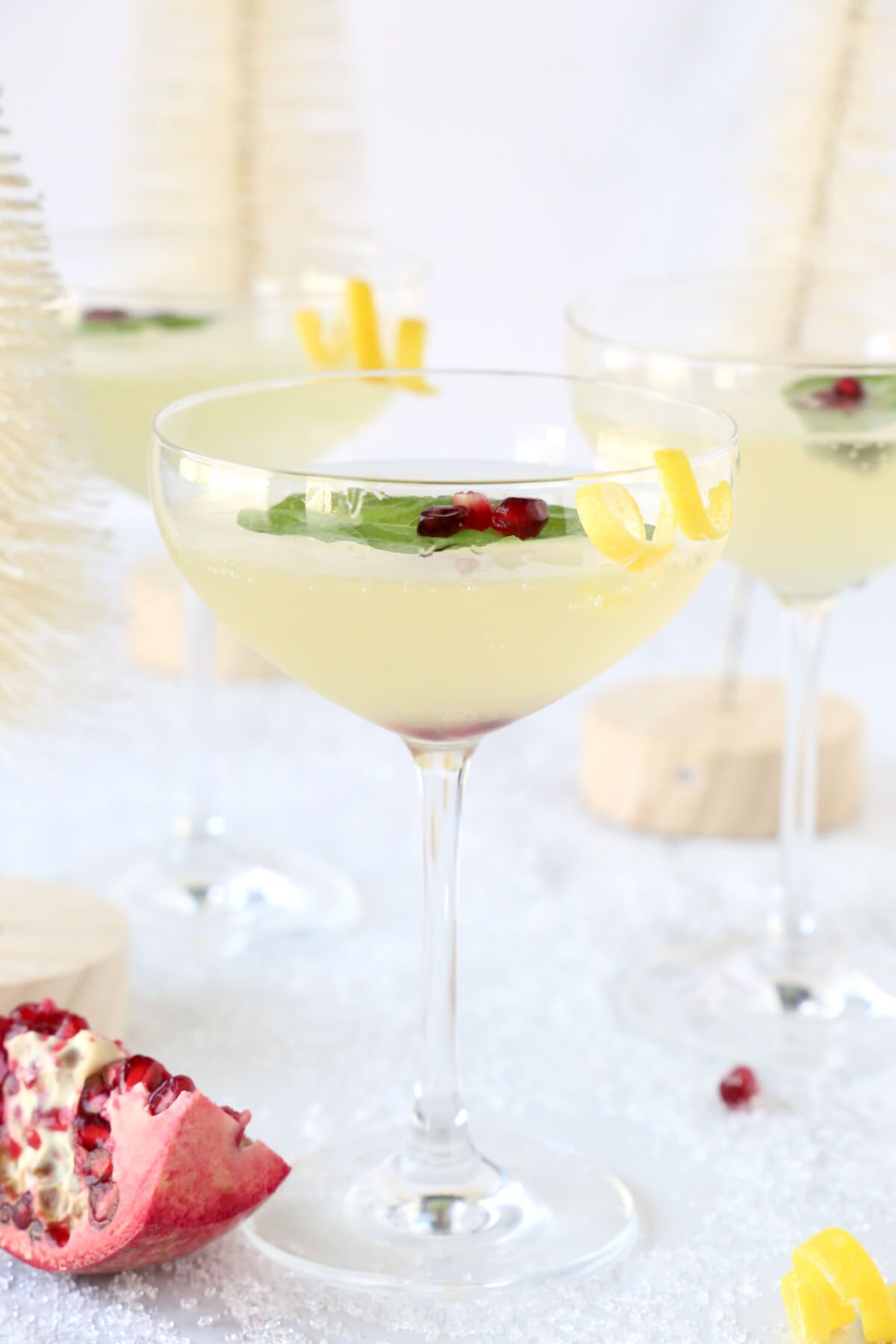 Three coupe glasses filled with a clear liquid, basil leaf, pomegranate seeds and a lemon rind curl.