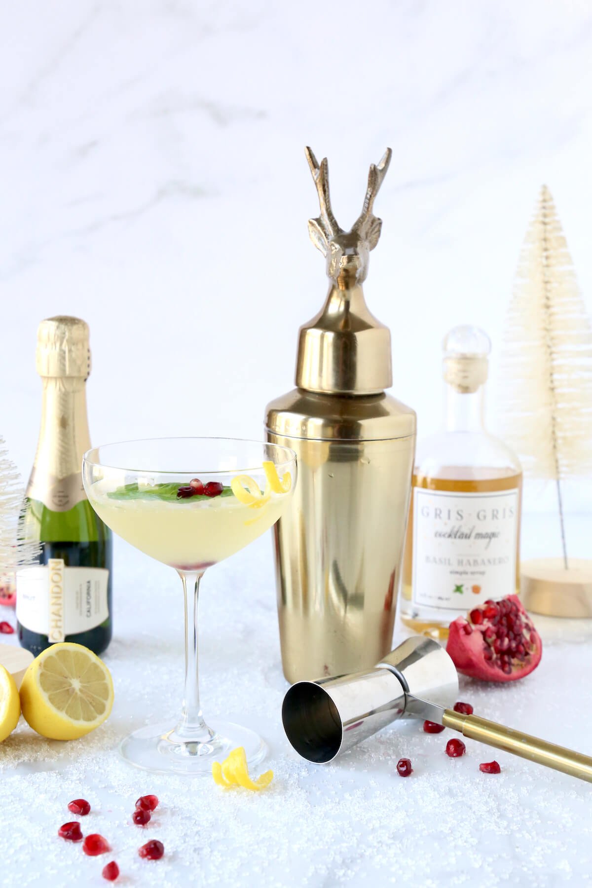 A gold cocktail shaker next to a small bottle of champagne, a glass jar, lemons, a glass with clear liquid and pomegranate seeds.