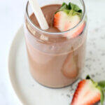A clear glass sitting on a white plate filled with chocolate brown smoothie, a white straw and a sliced strawberry.